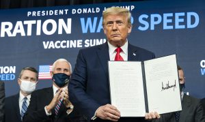 Donald Trump holding a signed document in front of a banner for Operation Warp Speed