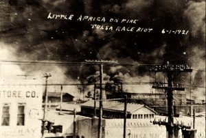 A sepia-tone photograph of smoke rising from buildings and hand-written text that reads, "Little Africa on fire; Tulsa Race Riot 6-/-/92/"