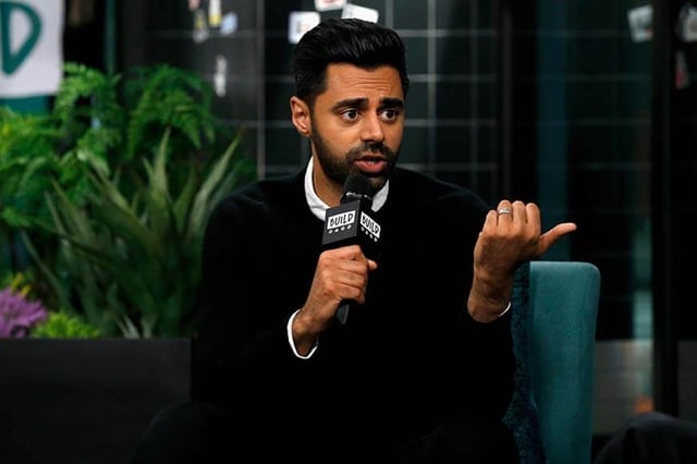 Comedian Hasan Minaj is shown speaking into a microphone.