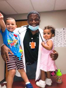 Chris McNeil wearing his white coat and stethoscope, smiling and holding his son Moses and daughter Madison