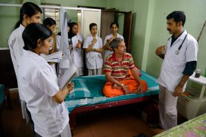 A Indian man sits on a medical bed surrounded by Indian doctors