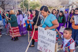 A group of Native Americans peacefully protesting. One woman and her child hold a sign that reads, "My mom, sisters, aunties, and grandmas are sacred."