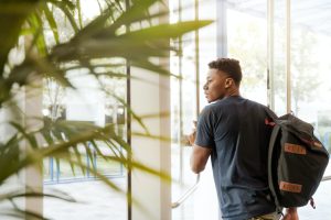 A black male student wearing a backpack and leaving a building through glass doors