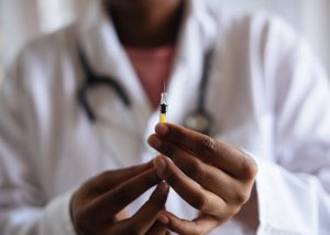 A person of color wearing a white coat and stethoscope and holding a vaccine